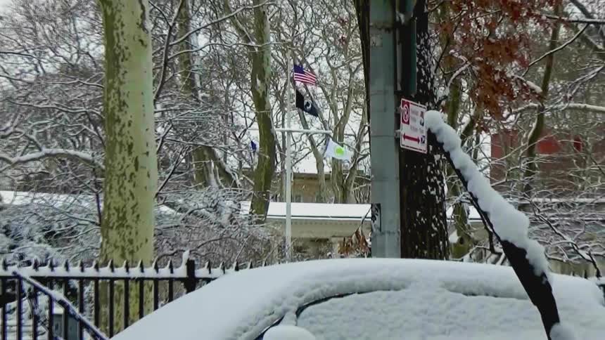 A shaky handheld camera pulls back to reveal a car covered in snow while across the street a person walks a dog through a quiet snowcovered park