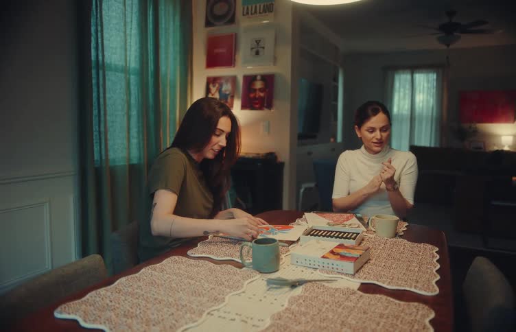 Two women sit at a dining table engaged in conversation while working on a craft project togetherthe room is warmly lit featuring artistic vinyl records on the wall and a cozy living space in the background