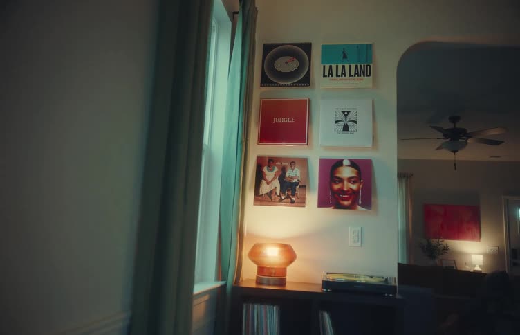 A living room at midday with a shelf beside the window a lit table lamp on top a spinning vinyl player beside it and several vinyl records hanging on the walls