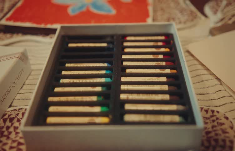 A womans hand reaches into a box of sennelier oil pastels picking one up as she works on a piece of cardboard laid out on the table
