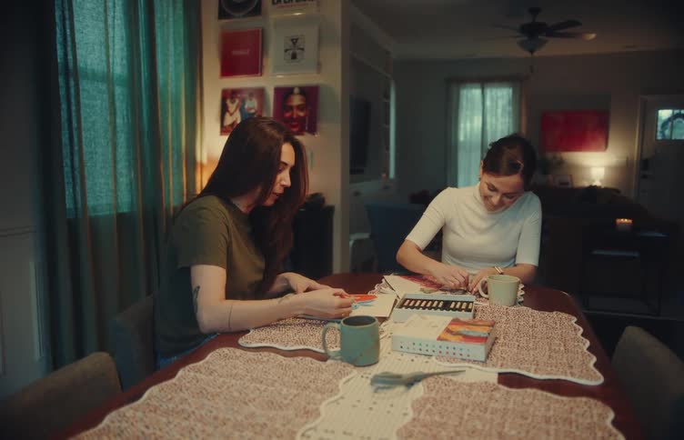 Two women are sitting at a dining table together while working on art projects they are focused on their coloring and sketching surrounded by art supplies and coffee mugs in a dimly lit cozy room