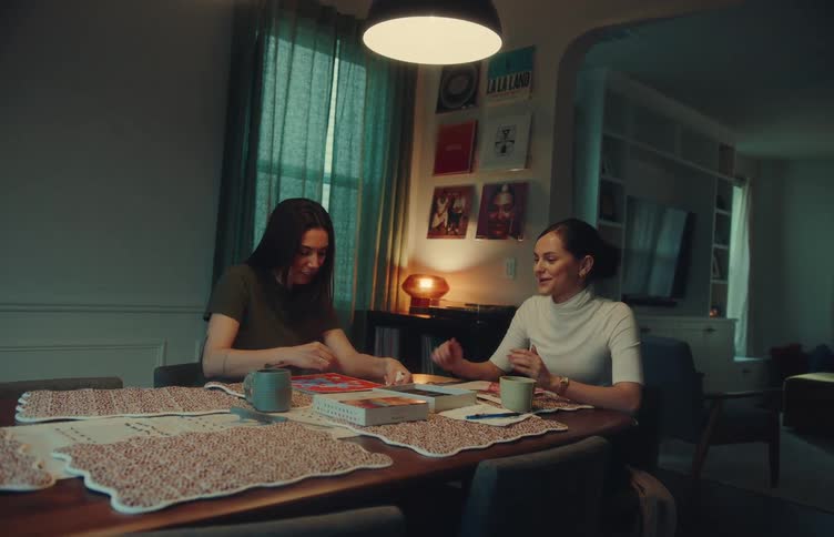 Two women sit at a wooden dining table under a hanging lamp while working on a jigsaw puzzle and talking together in a dimly lit room