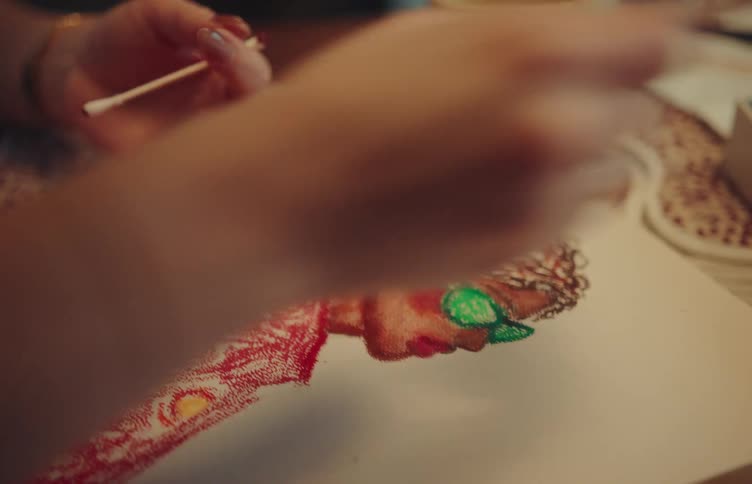 A close view shows the hands of a person using colorful crayons to draw a picture of a woman with large hair and green glasses on a white piece of paper