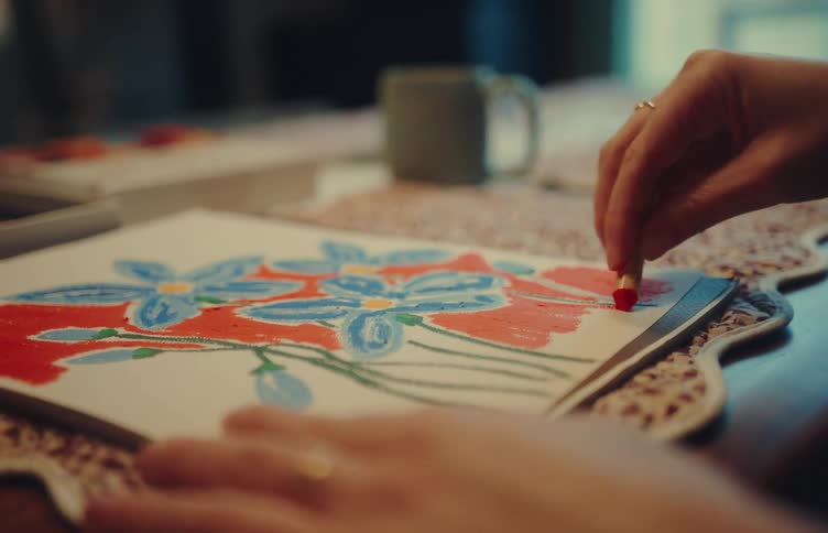 A person is using a red oil pastel to fill in the background around blue flower drawings on a white canvas the closeup shot focuses on the movement of their hand as they apply vibrant color in a cozy indoor setting