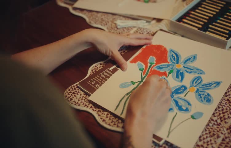 An artist crafts a colorful drawing in a sketchbook using vibrant oil pastels on a wooden table