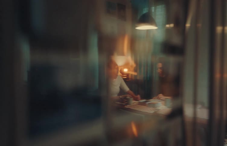 Two people are sitting at a table together in a dimly lit room appearing to be deeply engaged in a collaborative project or discussion the scene is shot through a glass surfacelikely a window or a glass doorwhich creates soft reflections and a sense o