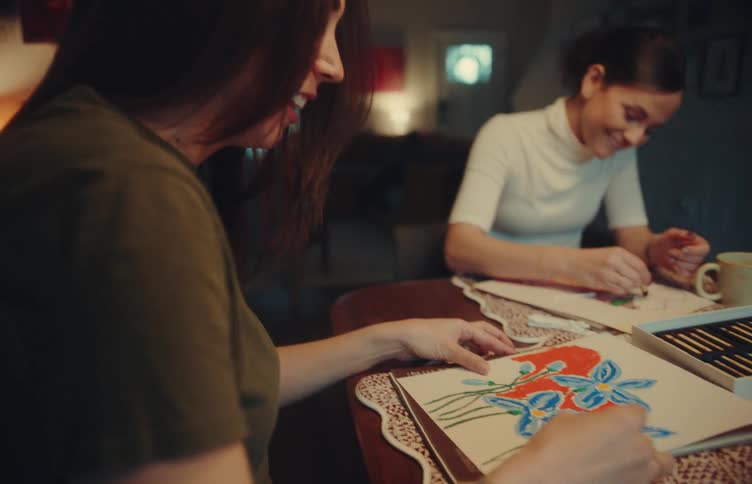 A close view shows two women sitting at a table while one uses colorful crayons to draw a picture of blue flowers on a white piece of paper as they talk and smile together