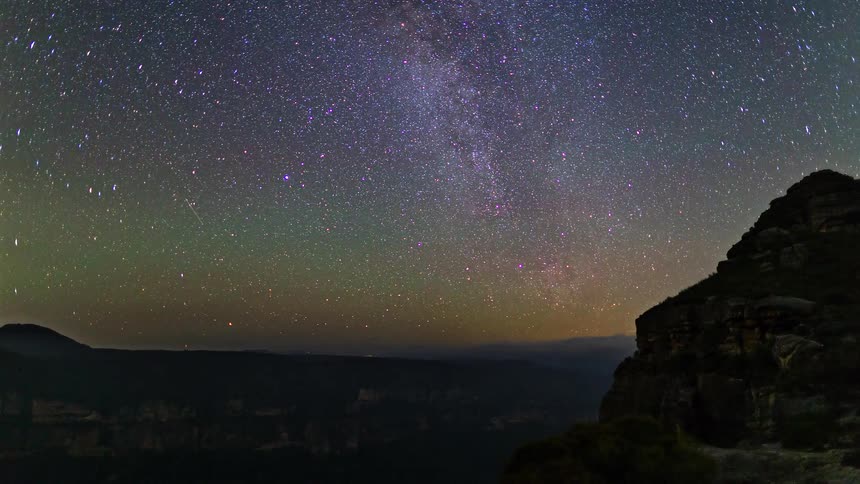 A wide night landscape with the milky way arching over starry skies and dark mountain silhouettes