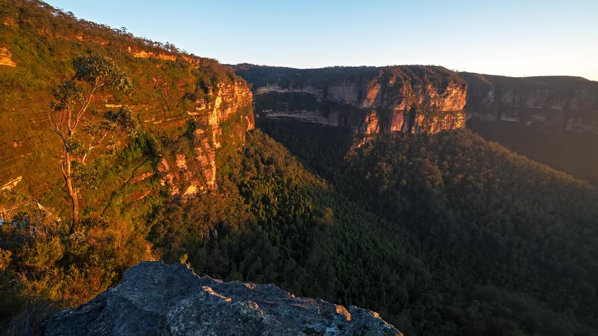 A view from a rocky cliff shows a deep valley filled with green trees and surrounded by tall orange rock walls under a clear blue sky during a bright sunrise