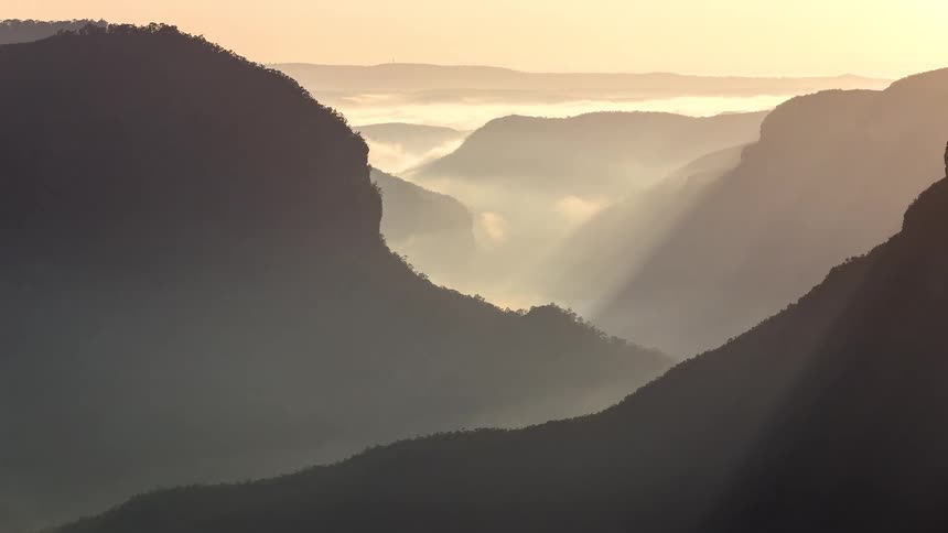 Sunbeams cut through the morning mist between silhouetted mountain ridges creating a soft golden glow across the valleythick clouds settle deep within the canyon floor as the early light reveals the layered depths of the rugged landscape