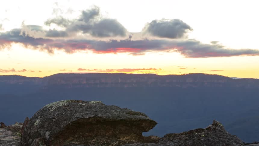 A rocky cliff overlooks distant mountains beneath dramatic clouds glowing at sunset