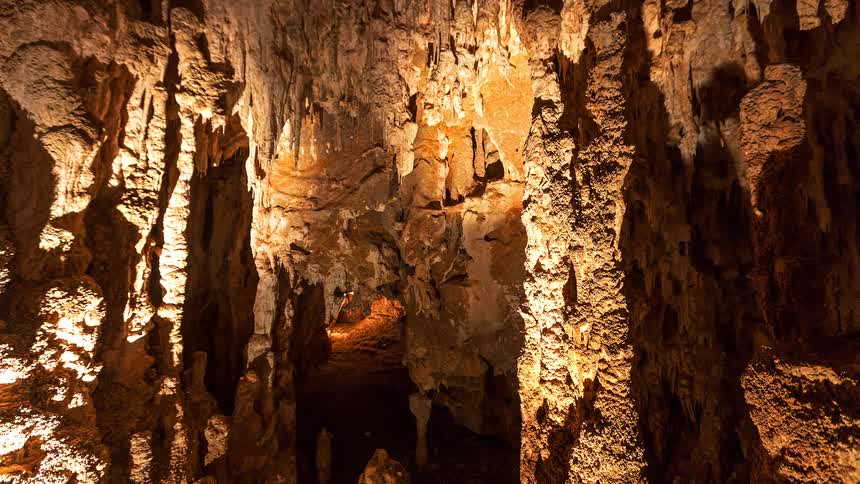 Warm orange light illuminates the intricate textures of stalactites and stalagmites within a deep limestone cavernthe camera slowly pans across the ancient rock formations revealing a narrow mysterious path leading further into the dark cave system