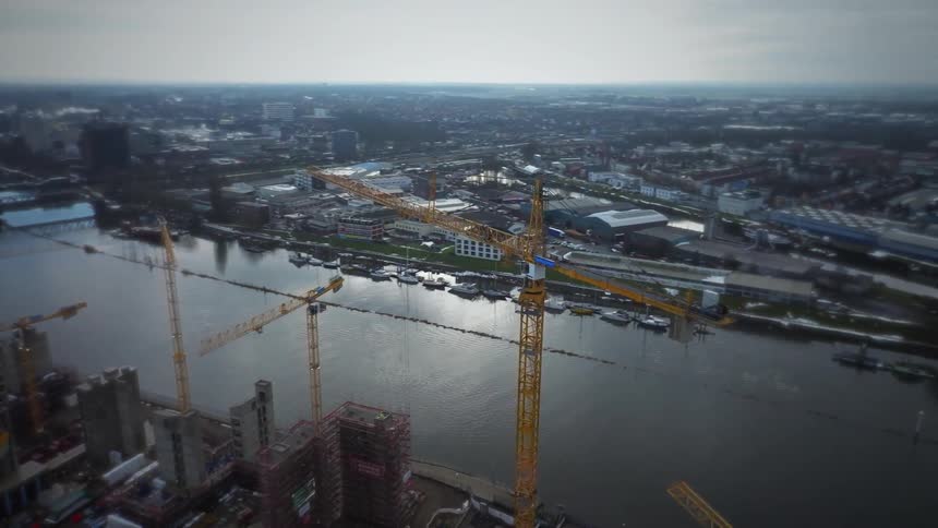 An aerial view shows several tall yellow construction cranes over a building site next to a wide river in a large city under a cloudy sky