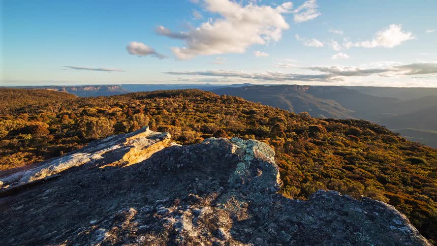 A view from a rocky cliff shows a landscape of mountains covered in green and orange trees under a blue sky with white clouds during a sunny day