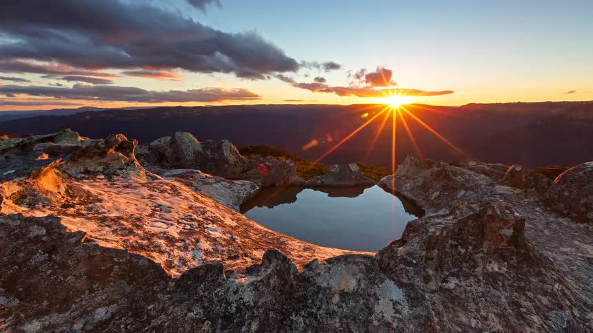 Sunlight illuminates a rocky mountain landscape with distant peaks and scattered clouds under a clear sky