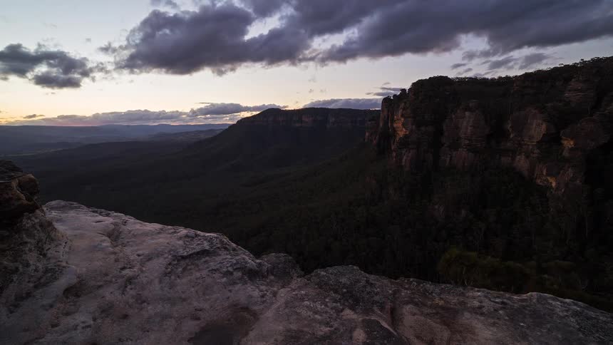 This timelapse video captures a golden sunset fading into twilight over a vast mountain valley and rocky cliffsas the sun disappears behind the horizon the vibrant orange light transitions into deep blue and purple hues across the darkening sky