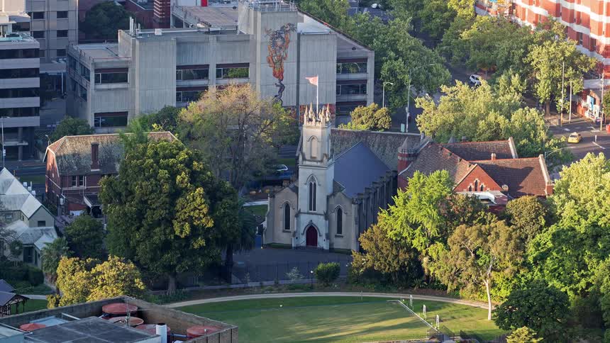 An aerial view shows a church with a tall tower and a red door while surrounded by many green trees and buildings in a city during the day