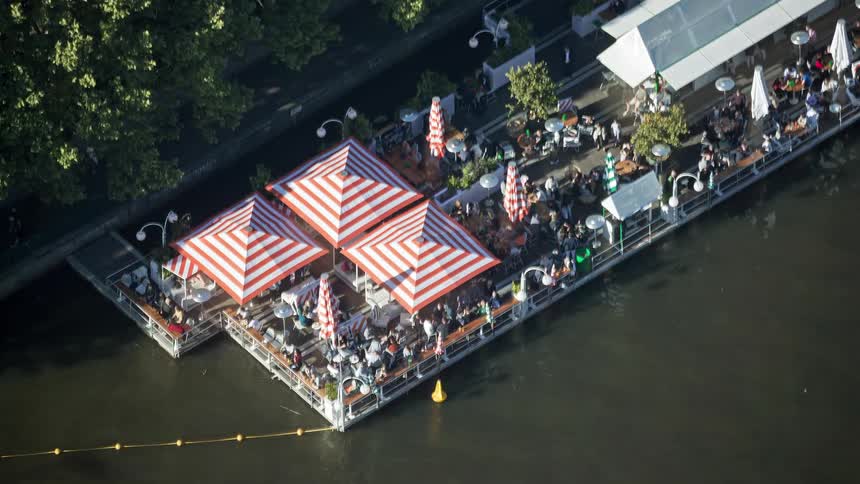 Crowd of people walking around or sitting down at a pier restaurant over a river next to a walkway on the shoreline outdoors during day time