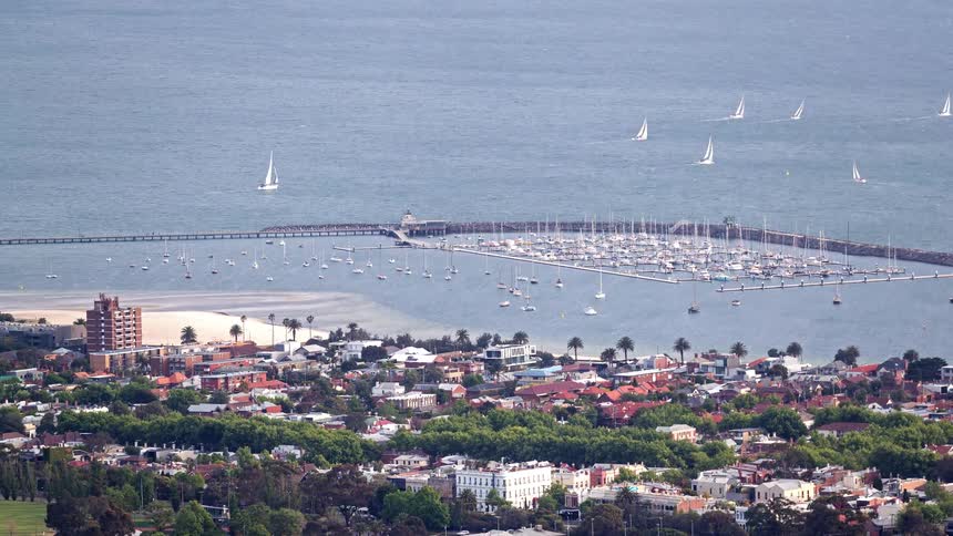 Marina with anchored sailboats and a coastal city in the background seen from above