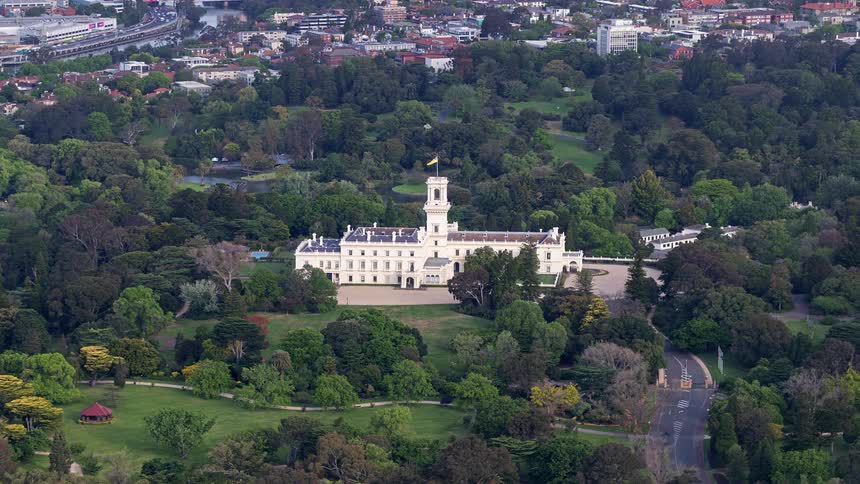 An aerial view of government house in melbourne a large white nineteenthcentury mansion with a prominent central tower it is surrounded by the lush greenery of the royal botanic gardens and the kings domain under a clear sky