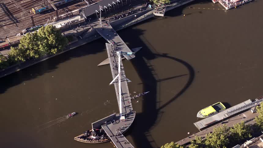 This aerial footage captures the evan walker bridge spanning the yarra river in melbourne featuring its distinct angular design and the long shadow it casts on the water rowing crews and small boats are seen gliding beneath the pedestrian walkway as peo