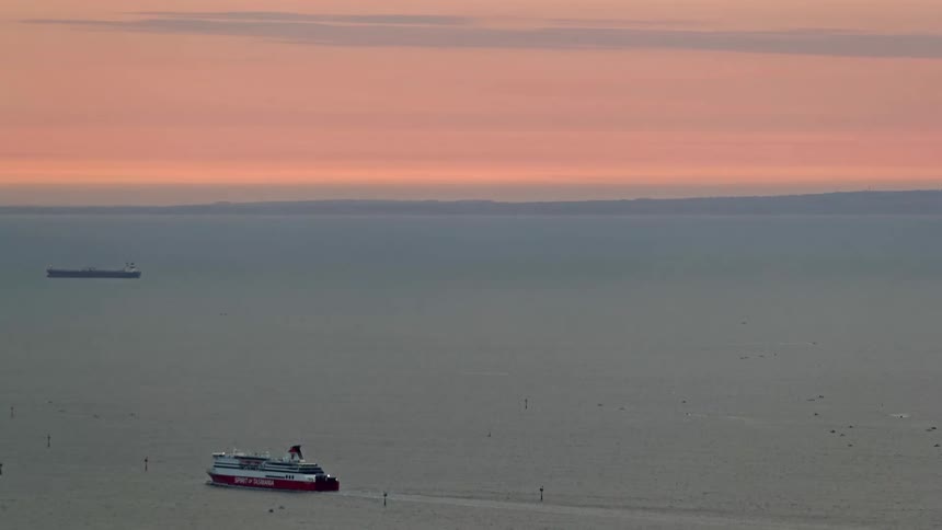 This video features a highangle shot of a spirit of tasmania ferry crossing a calm sea under a soft orangetinted sunset sky a large cargo ship sits stationary in the distance silhouetted against the hazy coastline