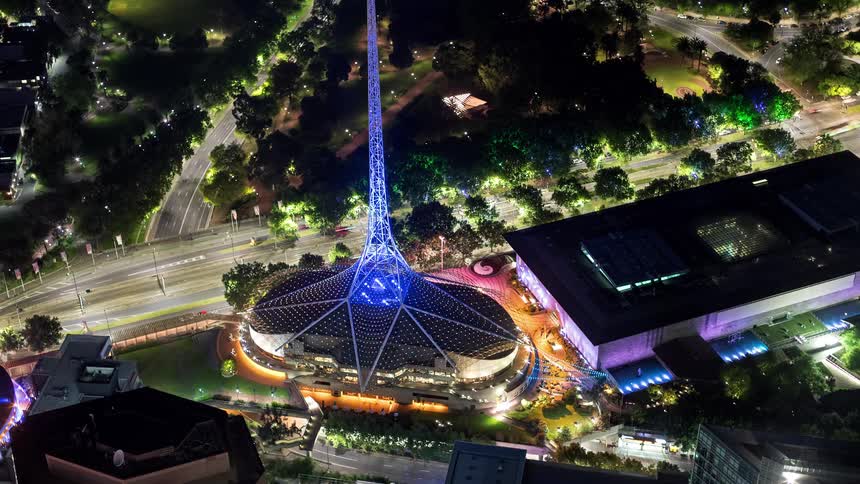 A stunning nighttime aerial view of the arts centre melbourne featuring its iconic illuminated spire and glowing mesh structure the surrounding city landscape is visible with bright streetlights lush green parks and the dark silhouettes of nearby buil