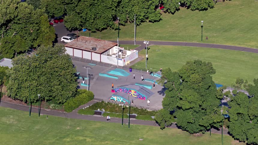 An aerial view shows several people skating at a park with blue ramps and colorful graffiti surrounded by green grass and trees during the day