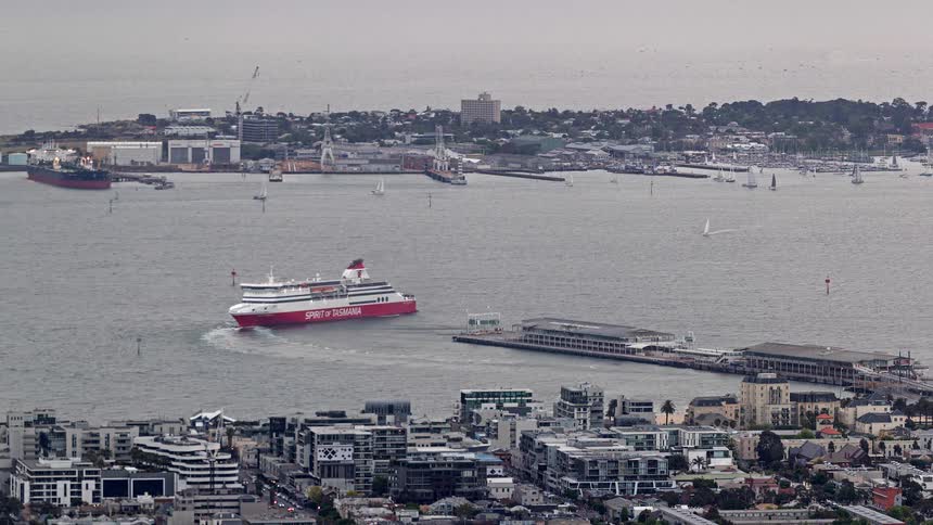 A passenger ferry moves across a busy harbor with a coastal city and port infrastructure in the background