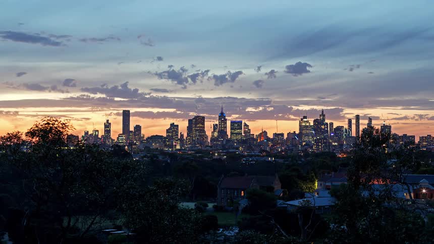 A view shows a city skyline at dusk with many buildings and houses as the sun sets and the sky turns orange and blue