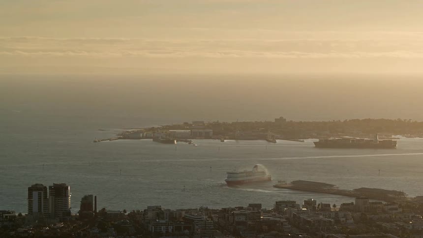 A large passenger ferry and a massive cargo ship navigate through the golden waters of a bay at dusk while a coastal city skyline sits in the foreground under a hazy horizon
