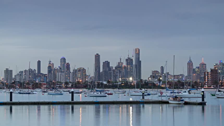 A view of a city skyline at dusk with many boats and yachts in the water as the lights of the buildings begin to turn on