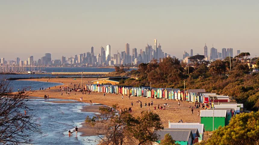 People on a beach full of colored cabins next to a grove with a metropolitan city on the background outdoors during different times of the day
