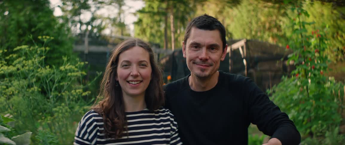 Young adult man and woman smiling on a yard with trees and a wooden shed on the background outdoors during day time