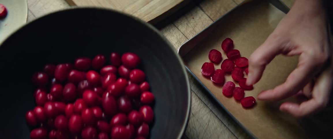 A person is placing chopped fresh red cranberries onto a baking sheet beside a black bowl also filled with berries all on a wooden surface