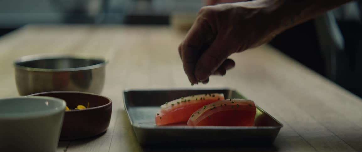 A close view shows the hands of a person as they sprinkle small green herbs onto two pieces of raw pink fish on a silver tray