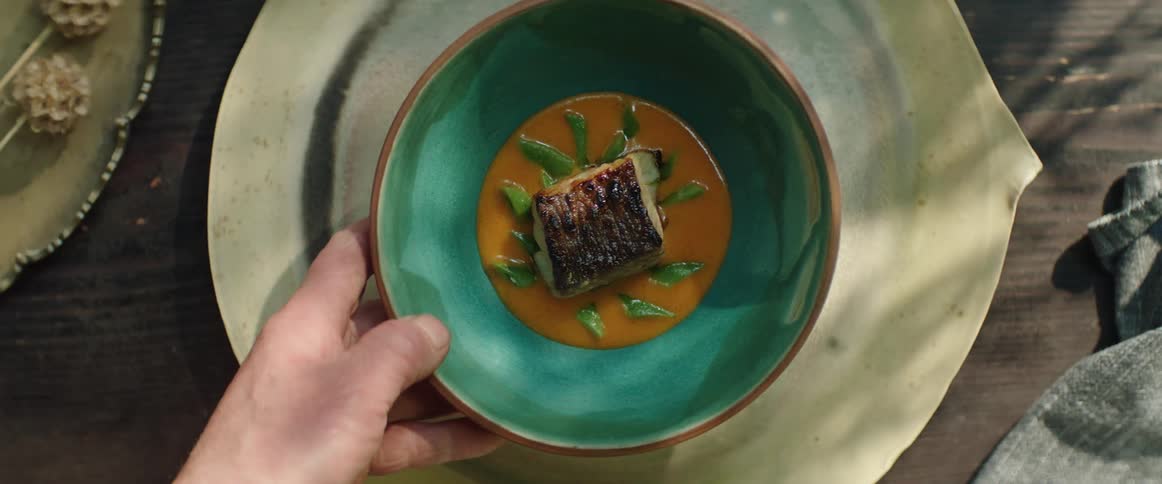 A hand removes a teal bowl containing seared fish from a large rustic plate