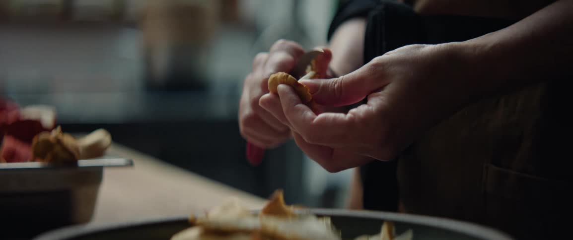 A close view shows the hands of a person as they use a small knife to trim the stem of an orange mushroom above a bowl in a kitchen