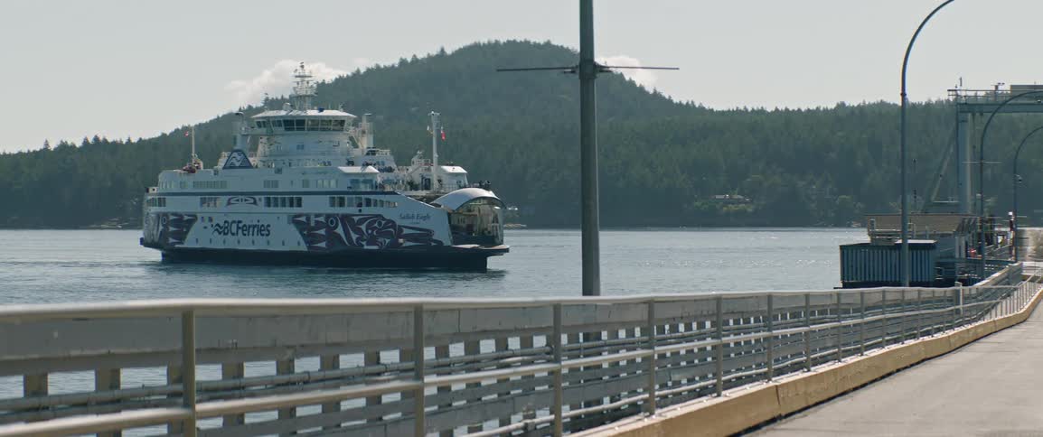 Videoa view from a dock shows a large ferry with blue and white markings as it moves across the water toward a shore covered in green trees during the day
