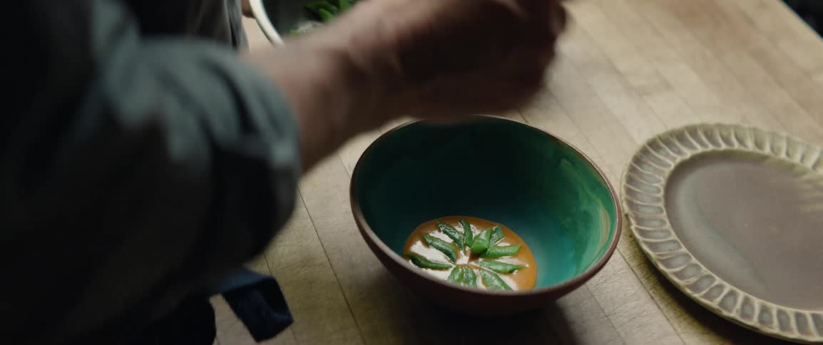 A person wearing a grey shirt and blue apron carefully places green snap peas into a bowl of orange soup with a spoon to create a floral pattern on a wooden countertop