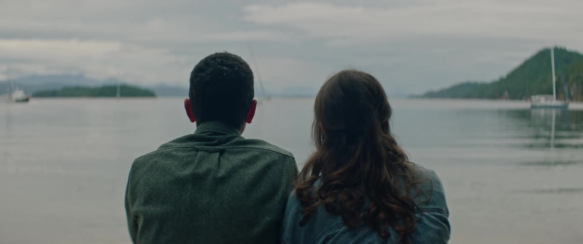 A couple sits together quietly gazing over calm water toward distant forested islands
