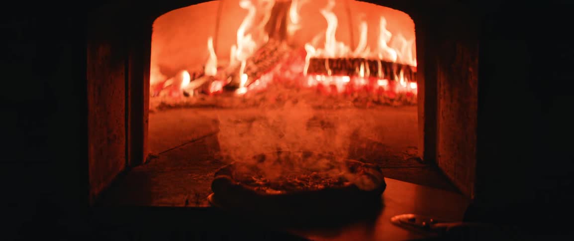 Pizza being removed from a woodfired oven with flames in the background