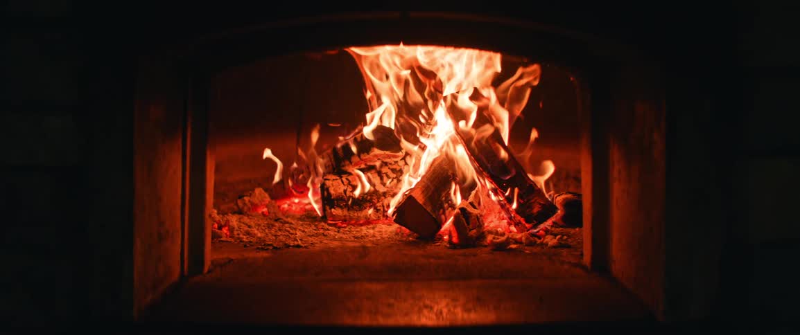 Bright orange flames lick the ceiling of a stone oven while thick logs burn steadily on a bed of glowing embers and gray ash inside a dark chamber