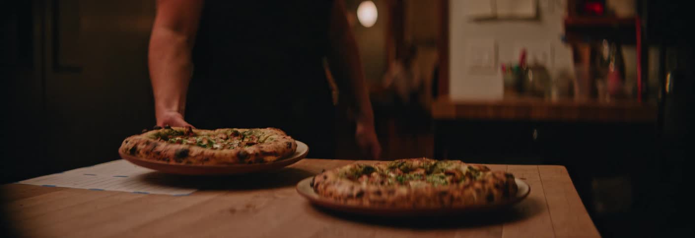 A man wearing a black apron reaches down to pick up a plate with a charred crust pizza from a wooden table while another pizza remains in the foreground of a dimly lit kitchen setting