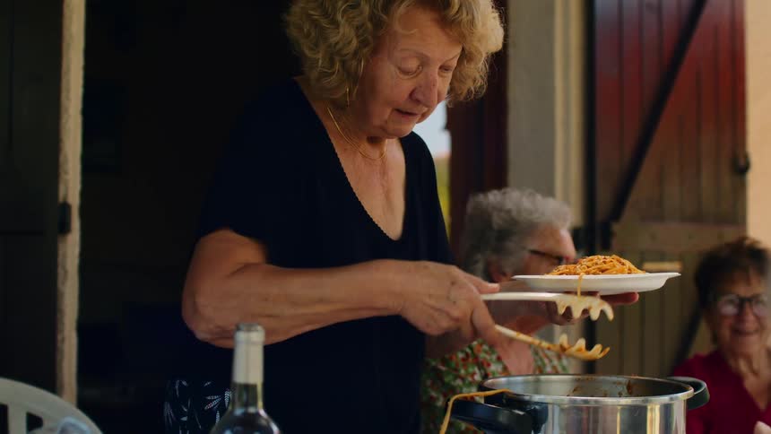 An elderly woman is serving spaghetti to several people