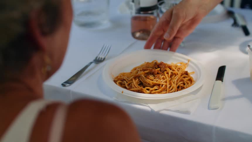 Adult male arm serving a dish of spaghetti over a an arranged table to an adult woman sitting and talking in front of it outdoors during day time