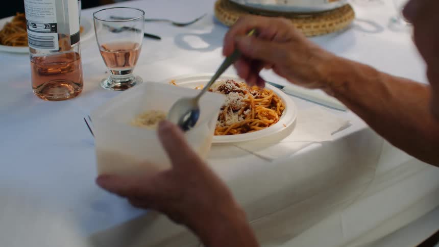 An elderly person uses a spoon to sprinkle grated cheese from a plastic container onto a plate of spaghetti with red sauce during a meal at a table covered in a white cloth with wine and glasses nearby