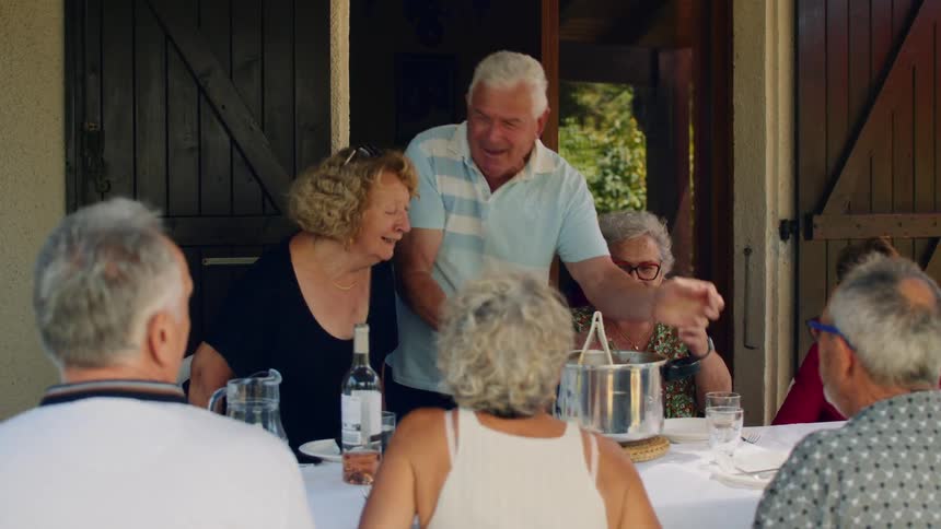 Elderly man carrying a sauce pan to a dinner table where other elderly people are sitting and talking outdoors during day time