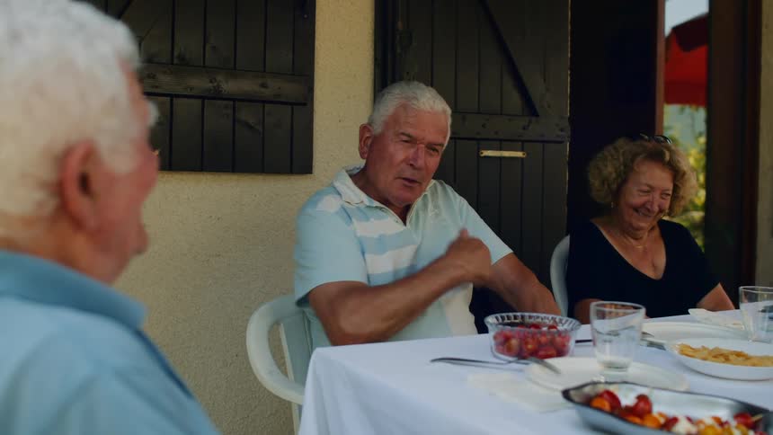 Three elderly people two men and one woman sitting at an outdoor dining table having lunch and smiling while talking