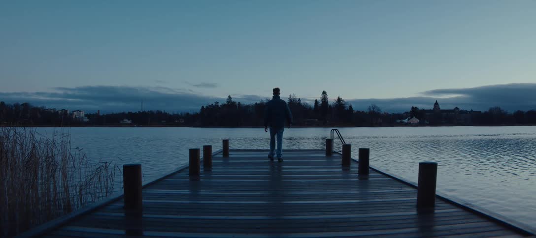 A view from behind shows a person walking toward the end of a wooden dock on a calm lake during the twilight hours